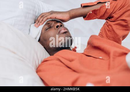 Der junge Afro-amerikanische Mann in einem orangefarbenen Hemd mit Aufnäher auf der Stirn leidet unter hohen Temperaturen, die in einem großen Bett in der Nähe der Seite liegen Stockfoto