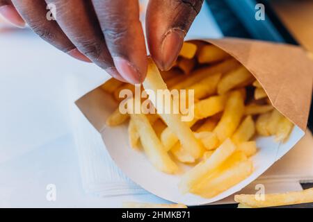 Ein junger afroamerikanischer Mann nimmt leckere frische pommes aus dem Papierbehälter an den Tisch im modernen Café, ganz aus der Nähe Stockfoto