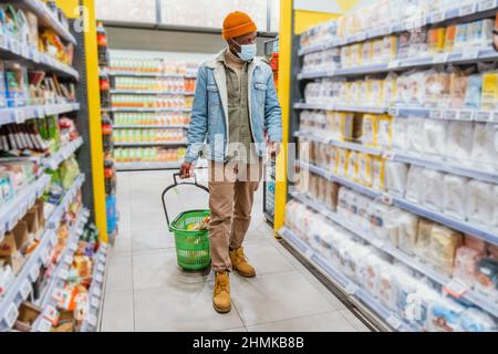Junger afroamerikanischer Mann in orangefarbenem Hut mit Schutzmaske geht und schaut auf Regale mit Waren in der Supermarktabteilung Stockfoto