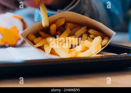 Ein junger afroamerikanischer Mann nimmt leckere frische pommes aus dem Papierbehälter an den Tisch im modernen Café, ganz aus der Nähe Stockfoto
