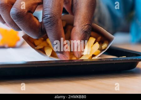 Ein junger afroamerikanischer Mann nimmt leckere frische pommes aus dem Papierbehälter an den Tisch im modernen Café, ganz aus der Nähe Stockfoto