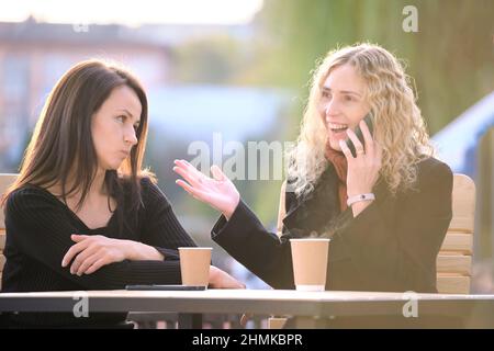 Glückliche junge Frau, die auf dem Handy chattet und ihre gelangweilte Freundin ignoriert. Weibliche Freunde sitzen im Straßencafe im Freien und haben harte Zeit zu kommunizieren Stockfoto