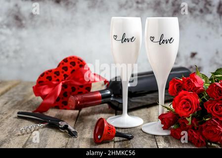 Valentinstag-Konzept. Zwei Gläser Wein, rote Rosen, Geschenk, Glocke, Weinflasche und Korkenzieher auf einem rustikalen Holztisch. Selektiver Fokus. Stockfoto