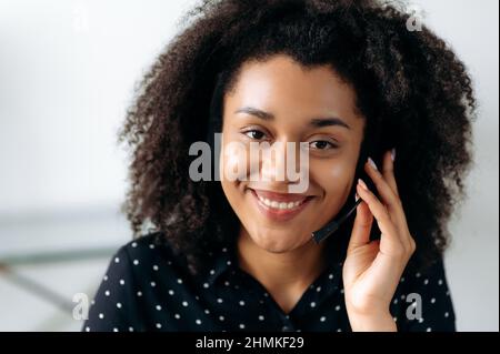 Nahaufnahme einer angenehmen, selbstbewussten afroamerikanischen Frau mit Kopfhörern, Freiberufler, Callcenter-Mitarbeiter, Berater, Telefonzentrale, In formellem Hemd, direkt auf die Kamera blickend, freundlich lächelnd Stockfoto