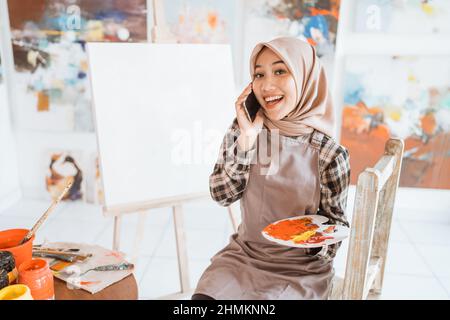 muslimische asiatische Künstlerin malen auf Leinwand und sprechen mit dem Telefon Stockfoto