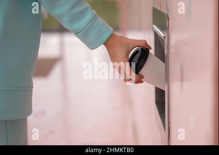Eine Frau wirft eine Tasse Kaffee in einen Mülltonnen in der Wand eines Cafés. Stockfoto