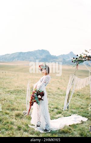 Die Braut in einem Kranz mit Blumenstrauß steht auf einem weißen Teppich in der Nähe eines Hochzeitsbogens Stockfoto