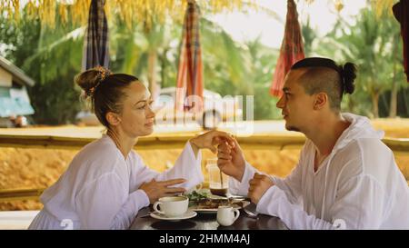 Attraktive junge Männer und Frauen essen und trinken leckere Getränke und verbringen Zeit in einem traditionellen, schwimmenden Café auf dem Wasser. Liebevolles glückliches Paar beim Frühstück im Freien, füttern sich gegenseitig. Tropischer Blick Stockfoto