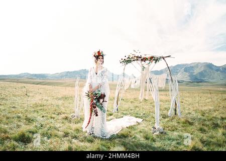 Braut in einem Kranz mit einem Strauß steht neben einem Hochzeitbogen im Rasen Stockfoto