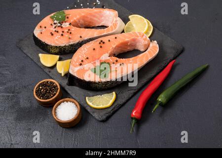 Zwei rohe Lachssteaks mit Gewürzen, Zitronenscheiben auf einem Steinbrett und schwarzem Hintergrund. Das Konzept, eine gesunde, vitaminreiche Mahlzeit zu kochen Stockfoto