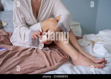 Die Frau sitzt im Bett im Bademantel und versorgt ihre Beine mit pflegender Creme. Das Konzept der Fußhautpflege zu Hause. Verjüngung und Ernährung der Haut des Körpers. Stockfoto