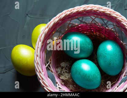 Bunt bemalte, glänzende Ostereier aus Marmor in einem Korb mit zarten Federn aus der Nähe. Nest mit Stroh - Osterdekoration auf dem Tisch Stockfoto