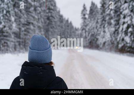 Frau Reisende Blick in die Ferne auf sich bewegende Auto, während auf verschneite Straße im Winterwald stehen Stockfoto