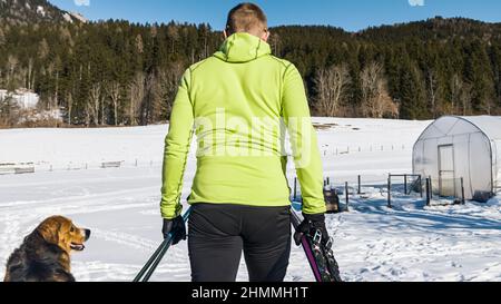 Ein Mann mittleren Alters aus dem Kaukasus in Skibekleidung, der mit seinem Hund durch den Schnee läuft und Langlaufskier und Skistöcke in den Händen hält. Stockfoto