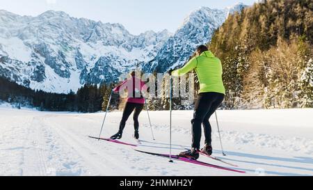 Kaukasisches Paar bewegt sich synchron während des Skilanglaufs auf der Skipiste, umgeben von Bergen und Wald. Stockfoto
