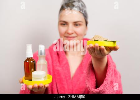 Eine Frau mit einem Film auf dem Kopf macht eine Haarmaske und hält natürliche und unnatürliche Kosmetik in den Händen Stockfoto