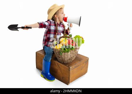 Ein süßes kleines Mädchen, emotioniges Kind im Bild des Bauern, Gärtner mit großem Korb mit Gemüse isoliert auf weißem Hintergrund. Konzept von Job, Arbeit Stockfoto