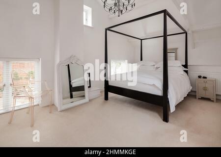 Newport, Essex - Juli 10 2018: Luxuriöses, modern eingerichtetes, schickes Schlafzimmer in Weiß mit Himmelbett aus dunklem Holz und einem auffälligen Spiegel mit kunstvollem Rahmen Stockfoto