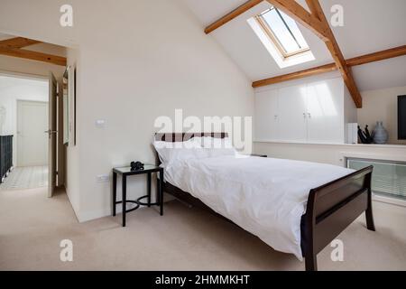 Newport, Essex - Juli 10 2018: Modisches, minimalistisches, weiß gestrichenes, sonnenüberflutete Schlafzimmer mit Bett, Beistelltisch und gewölbter Decke mit freiliegenden Balken. Stockfoto