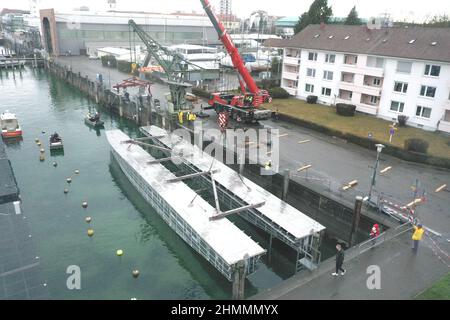 Friedrichshafen, Deutschland. 11th. Februar 2022. Zwei Krane heben die beiden Rümpfe für den zukünftigen E-Katamaran von Land in das Hafenbecken. (Luftaufnahme mit einer Drohne) die Rümpfe wiegen rund 20 Tonnen und wurden in Stralsund gebaut. Am Morgen kamen die Rümpfe von der Werft mit einem Schwerlasttransporter in Friedrichshafen an. Quelle: Felix Kästle/dpa/Alamy Live News Stockfoto