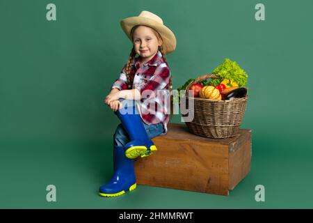 Porträt von niedlichen kleinen Mädchen, emotive Kind in Bild von Landwirt, Gärtner sitzt in der Nähe von riesigen Korb mit Gemüse isoliert auf grünem Hintergrund. Konzept Stockfoto