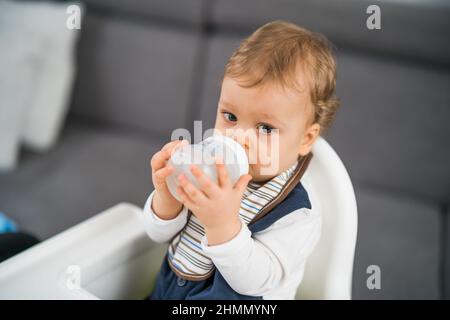 Netter kleiner Junge, der Wasser trinkt, während er auf einem Hochstuhl sitzt. Stockfoto