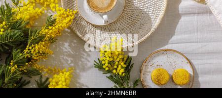 Zweige gelber Mimosenblüten auf weißen Leinenbettlaken, Tasse oder Kaffee, französische gelbe Makronen. Gruß zum Internationalen Frauentag am 8. März Stockfoto