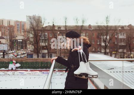 Die junge, lockige Frau in einer Baskenmütze mit Schlittschuhen blickt auf die Eisbahn herab, will lernen, wie man Schlittschuh laufen kann Stockfoto