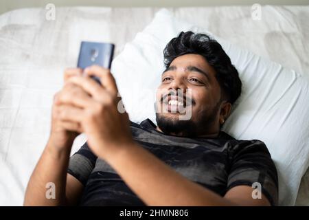 Glücklicher junger Mann, der auf dem Handy chattet, während er im Bett schläft - Konzept der Verwendung von Social Media, Dating-App, Flirten und Technologie. Stockfoto