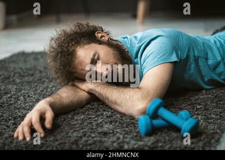 Erschöpft von Heimübung Mann lag auf dem Teppich und ruht sich aus. Lustige zerzauste Haar Mann tun Übung zu Hause. Müder junger Mann, der zu Hause in Quarantäne Übungen macht. Konzept eines gesunden Lebens. Stockfoto