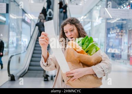 Eine Frau unter Schock blickt vor dem Hintergrund einer Rolltreppe in einen Papierscheck aus einem Supermarkt in einem Einkaufszentrum und hält ein Paket mit frischen Produkten, Preiserhöhung Stockfoto