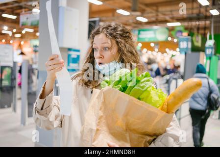 Eine junge Frau in einer medizinischen Maske sieht schockiert über einen Papierscheck in einem Supermarkt, der einen Papierbeutel mit Lebensmitteln, Preissteigerungen und Inflation hält Stockfoto