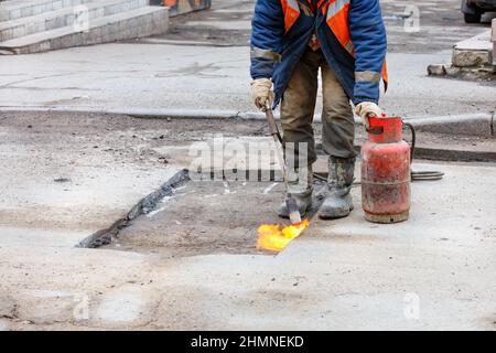 Ein Straßenarbeiter bereitet ein Schlagloch im Asphalt zur Reparatur vor, indem er den beschädigten Asphaltbelag mit einem Gasbrenner erhitzt. Stockfoto