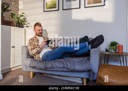 Eine Seitenansicht eines jungen Mannes, der entspannt auf seinem Sofa in seinem Wohnzimmer liegt und sein Smartphone benutzt. Stockfoto