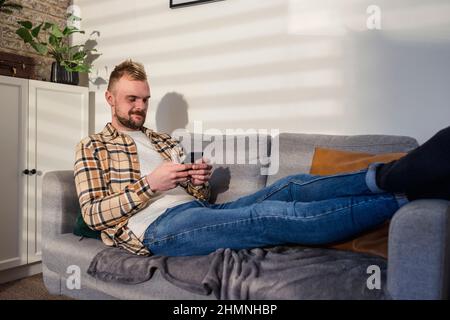 Eine Seitenansicht eines jungen Mannes, der entspannt auf seinem Sofa in seinem Wohnzimmer liegt und sein Smartphone benutzt. Stockfoto