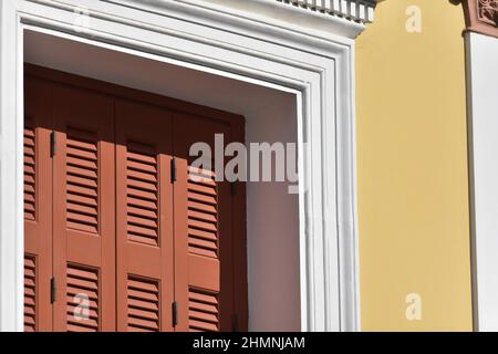 Altes neoklassizistisches Hausfenster mit hölzernen Fensterläden und einer weißen Zierleiste an einer ockerfarbenen Stuckwand in Nafplio, Griechenland. Stockfoto
