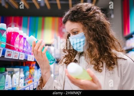 Porträt einer Frau Shopper mit lockigen Haaren trägt medizinische Maske im Supermarkt wählt Shampoo während Coronavirus Ausbruch Stockfoto