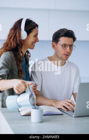 Lockig langhaarige Frau mit Kopfhörern lächelt und gießt Kaffee in den Becher für gestressten Ehemann, der am Laptop arbeitet und Arbeitsprobleme löst Stockfoto