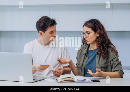 Ehemann und Ehefrau berechnen die Kosten der Finanzen sitzen zu Hause in der Küche am Tisch und schreiben Sie es in ein Notizbuch, eine Frau mit Schecks in den Händen macht Entschuldigungen, ein Mann ärgert sich Stockfoto