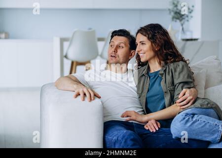 Glückliche Brautpaar mit dunklem Haar sitzen auf einem bequemen Sofa im Wohnzimmer und besprechen Pläne für ein Lächeln in der Zukunft ganz aus der Nähe Stockfoto