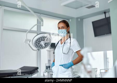 Porträt einer jungen Ärztin in medizinischer Maske und weißem Mantel, Stethoskop über dem Hals in der Klinik Stockfoto