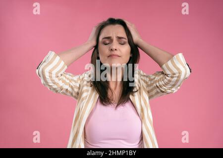 Gestresst oder mit Kopfschmerzen Geschäftsfrau in Anzug hält ihren Kopf mit geschlossenen Augen isoliert auf rosa Hintergrund. Gestresste Angestellte Frau im Streifenanzug. Mit Kopierbereich. Stockfoto