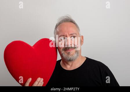 Schöner grauhaariger und bärtiger Mann mittleren Alters mit einem roten Herzen und isoliertem grauen Hintergrund. Valentinstag-Konzept Stockfoto