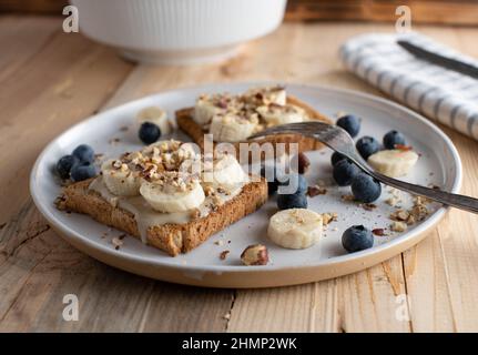 Energy Frühstücksteller mit Vollkorntoast, gekrönt mit Mandelbutter, Bananen und Haselnüssen. Serviert mit frischen Heidelbeeren isoliert auf Holztisch Stockfoto