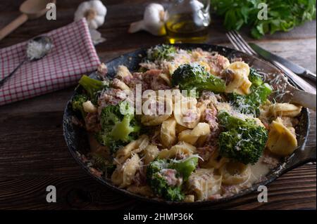 Tortellini mit Schinken, Parmesankäse und Brokkoli, serviert in einer rustikalen gusseisernen Pfanne auf einem Holztisch Stockfoto