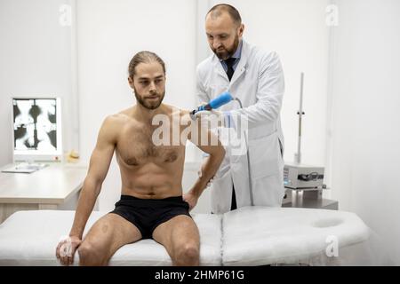 Der Arzt wendet eine Schockwellentherapie auf die Schulter des Mannes an Stockfoto