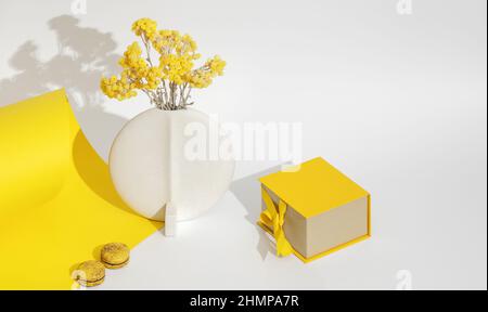 Unvergängliche, immortelle gelbe Blüten in minimalistischer weißer Keramikvase, zwei französischen Makronen und einer gelben Geschenkbox mit Schleife über Gelb und WH Stockfoto