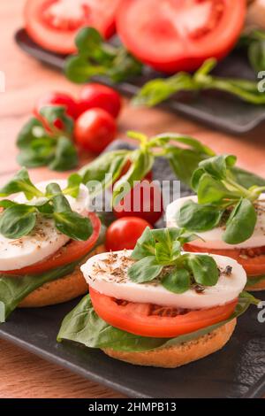 Vertikales Bild des klassischen italienischen Caprese-Salats mit geschnittenen Tomaten, Mozzarella und Basilikum auf Bruschettas auf schwarzem Teller auf Holztischhintergrund. T Stockfoto