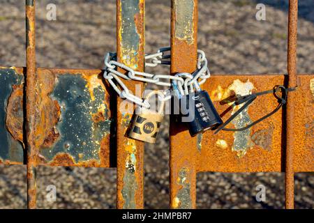 Nahaufnahme der Ketten und zwei Vorhängeschlösser, die ein Paar rostiger Metalltore in einer verlassenen Fabrik, einen Yale-Schlüssel und ein ASEC-Zahlenschloss sichern. Stockfoto