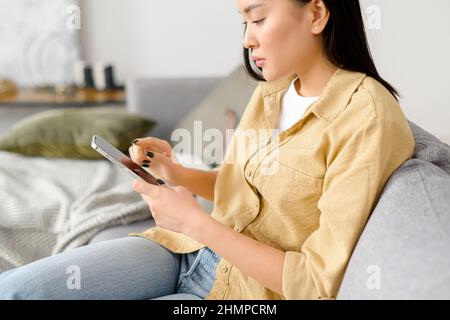 Seitenansicht der asiatischen Frau, die in ihren Händen auf das Smartphone schaut. Frau telefoniert, während sie sich auf dem Sofa sitzt. Technologien und Menschen Konzept Stockfoto